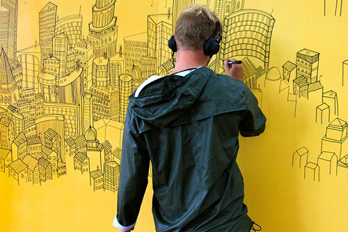 Man in a green jacket, wearing headphones, who is drawing a cityscape on a large yellow wall